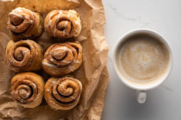 Vue de dessus des rouleaux de cannelle maison fraîche sur papier parchemin sur la surface de marbre avec café — Photo de stock