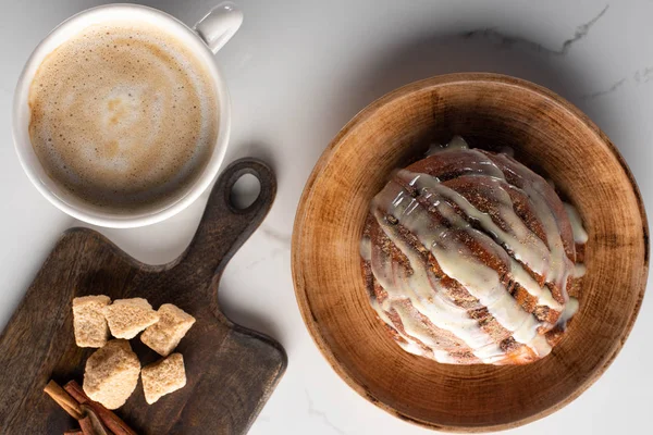 Vista dall'alto del fresco rotolo di cannella fatto in casa sul piatto sulla superficie di marmo con tagliere con zucchero di canna, bastoncini di cannella vicino al caffè — Foto stock