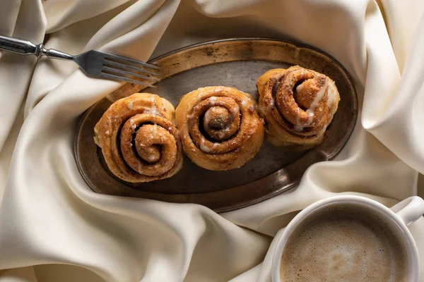 Top view of fresh homemade cinnamon rolls on board on satin cloth with fork and coffee — Stock Photo