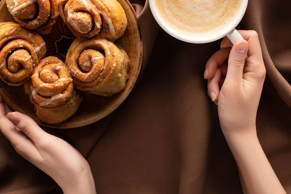 Vista ritagliata della donna che tiene il piatto con rotoli di cannella fatti in casa freschi vicino al caffè su un panno marrone seta — Foto stock