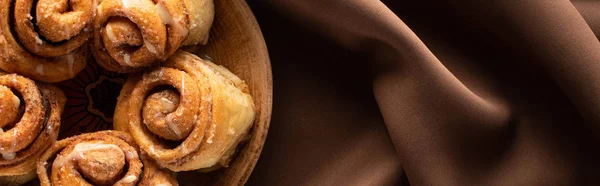 Top view of fresh homemade cinnamon rolls on plate on silk brown cloth, panoramic shot — Stock Photo