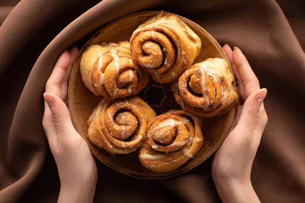 Cropped view of woman holding plate with fresh homemade cinnamon rolls on silk brown cloth — Stock Photo