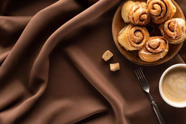 Top view of fresh homemade cinnamon rolls on silk brown cloth with brown sugar, fork and coffee — Stock Photo