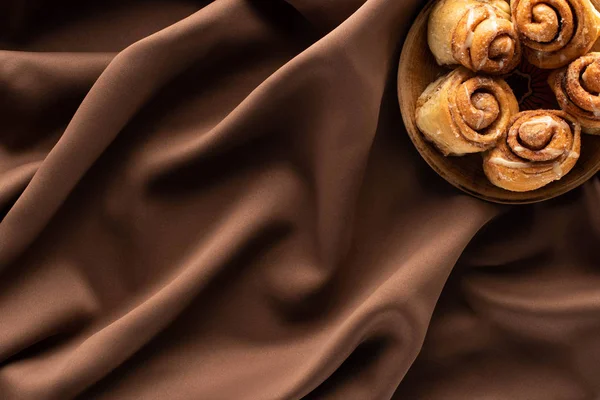 Vista superior de rollos de canela caseros frescos sobre tela marrón de seda - foto de stock