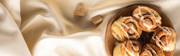 Top view of fresh homemade cinnamon rolls on silk white cloth with brown sugar, panoramic shot — Stock Photo