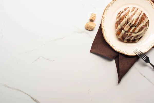 Top view of fresh homemade cinnamon roll on plate on marble surface with brown sugar, napkin and fork — Stock Photo