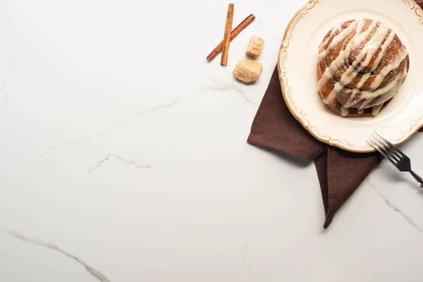 Top view of fresh homemade cinnamon roll on plate on marble surface with brown sugar, cinnamon sticks, napkin and fork — Stock Photo