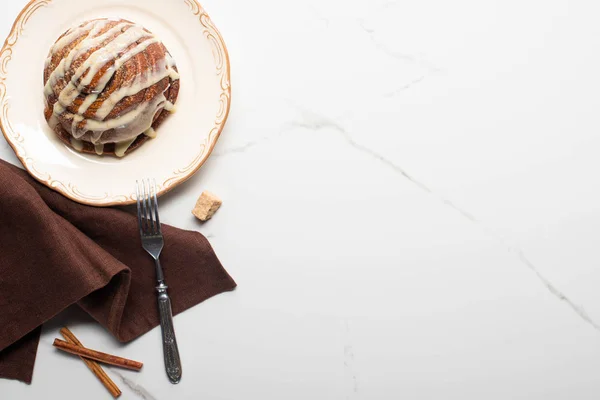 Draufsicht auf frische hausgemachte Zimtrolle auf Teller auf Marmoroberfläche mit braunem Zucker, Zimtstangen, Serviette und Gabel — Stockfoto