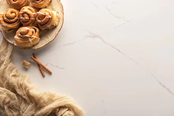 Vista superior de rollos de canela caseros frescos en la superficie de mármol con azúcar morena, palitos de canela y tela - foto de stock