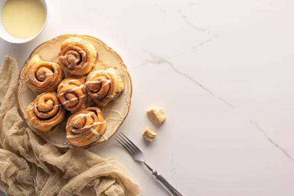 Top view of fresh homemade cinnamon rolls on marble surface with condensed milk, brown sugar, fork and cloth — Stock Photo