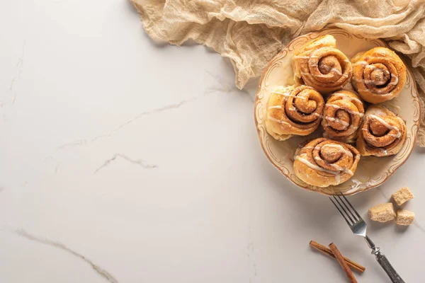 Vista superior de rollos de canela caseros frescos en la superficie de mármol con azúcar morena, palitos de canela, tenedor y tela — Stock Photo