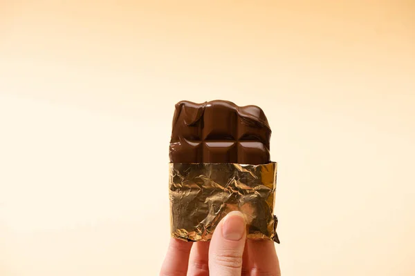 Cropped view of woman holding delicious milk chocolate bar in golden foil isolated on beige — Stock Photo