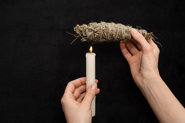 Cropped view of shaman holding herbal smudge stick above burning candle isolated on black — Stock Photo