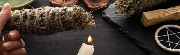 Cropped view of shaman holding smudge stick and candle near witchcraft on black wooden surface, panoramic shot — Stock Photo