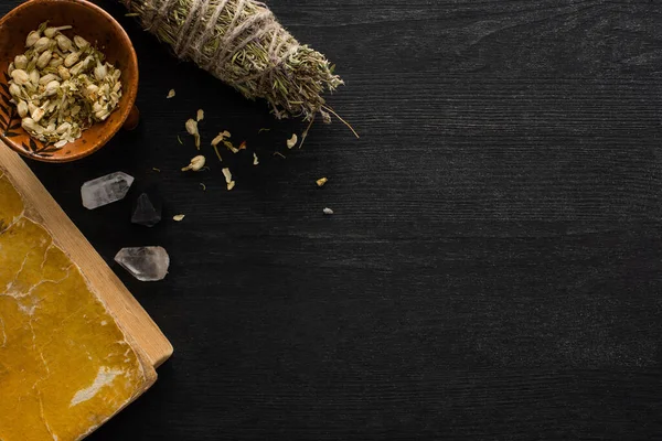 Vista dall'alto di bastoncini di sbavatura, fiori secchi in ciotola e libro sulla superficie in legno nero — Foto stock