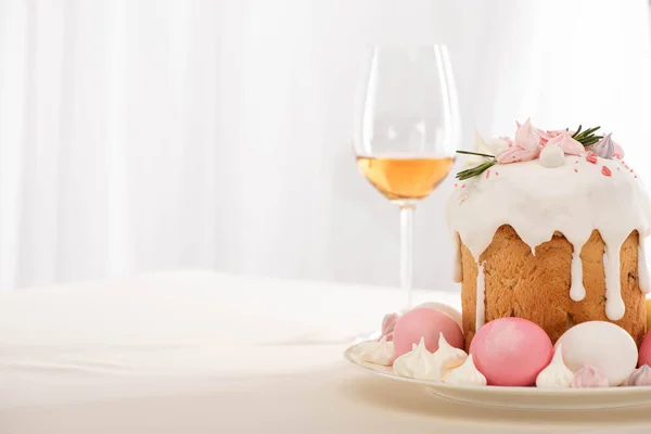 Вкусный пасхальный торт украшен безе с розовыми и белыми яйцами на тарелке рядом с бокалом вина — стоковое фото