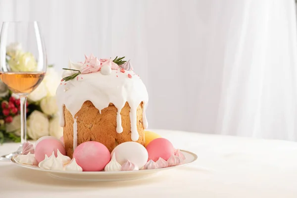 Селективный фокус вкусного пасхального торта, украшенного безе с розовыми и белыми яйцами на тарелке возле бокала вина и цветов — стоковое фото
