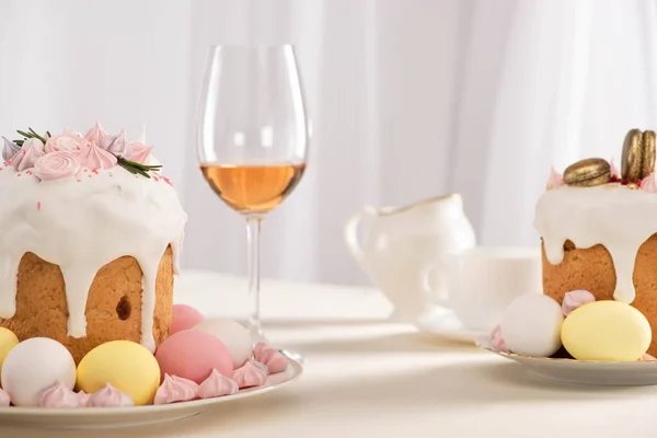 Delicious Easter cakes decorated with meringue near colorful eggs on plates and wine glass — Stock Photo