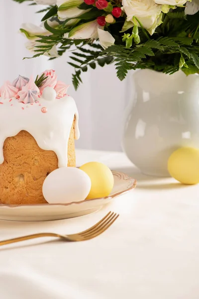 Delicioso bolo de Páscoa decorado com merengue perto de ovos coloridos na placa na mesa com vaso de flores e garfo — Fotografia de Stock