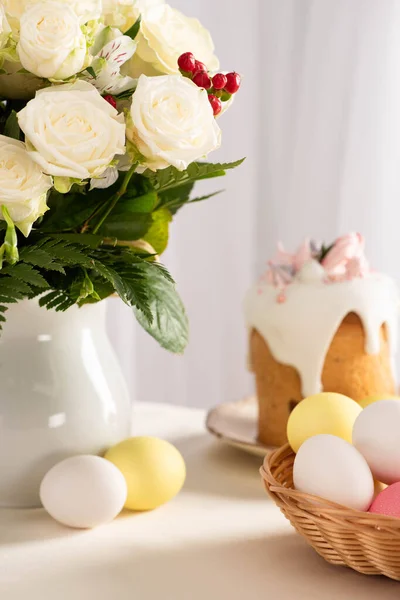 Селективный фокус праздничного пасхального торта, украшенного безе возле окрашенных яиц в корзину и вазу с цветами — стоковое фото