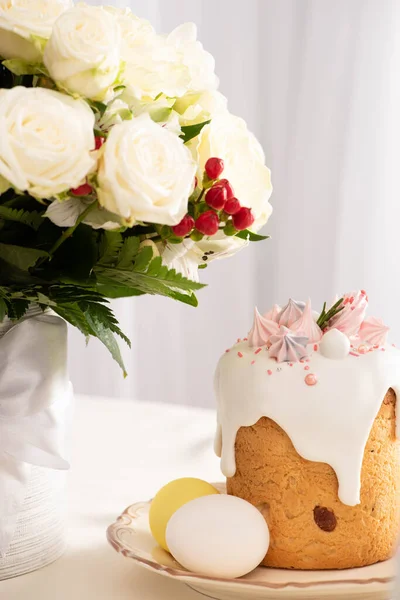 Festive Easter cake decorated with meringue near painted eggs on plate and vase of flowers — Stock Photo