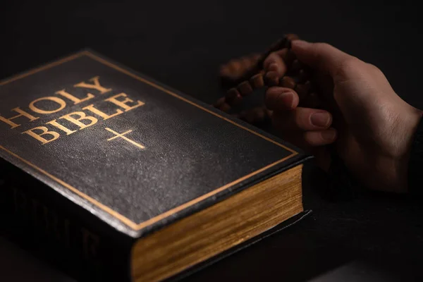 Cropped view of woman holding rosary near holy bible in dark with sunlight — Stock Photo