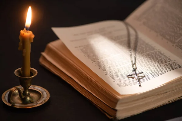 KYIV, UKRAINE - JANUARY 17, 2020: church candle in candlestick burning near bible with catholic cross in dark with sunlight — Stock Photo