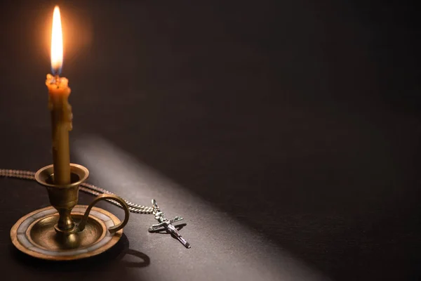 Church candle in candlestick burning near catholic cross in dark with sunlight — Stock Photo