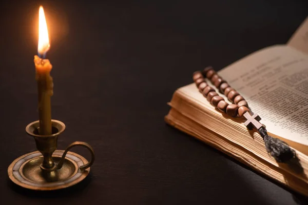 KYIV, UKRAINE - 17 JANVIER 2020 : bougie d'église au chandelier brûlant près de la bible avec chapelet catholique dans l'obscurité — Photo de stock