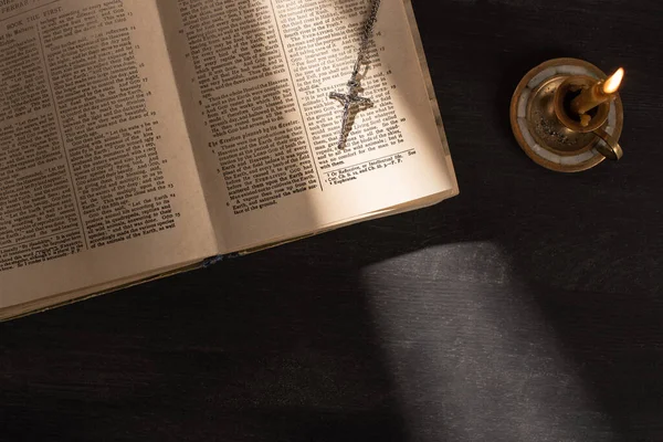 КИЕВ, УКРАИНА - 17 ЯНВАРЯ 2020 г.: вид сверху на открытую Святую Библию с крестом возле свечи на темном фоне с солнечным светом — стоковое фото