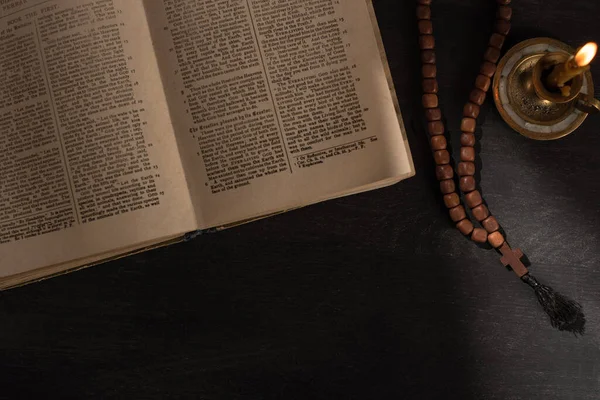 KYIV, UKRAINE - JANUARY 17, 2020: top view of open holy bible with rosary near candle on dark background with sunlight — Stock Photo