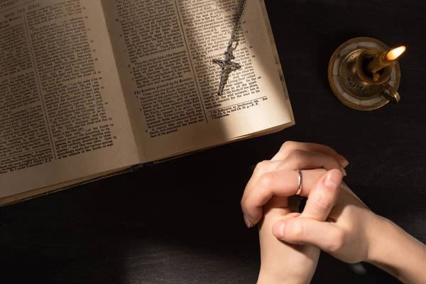 KYIV, UKRAINE - JANUARY 17, 2020: cropped view of woman praying near open holy bible with cross on dark background with sunlight — стокове фото