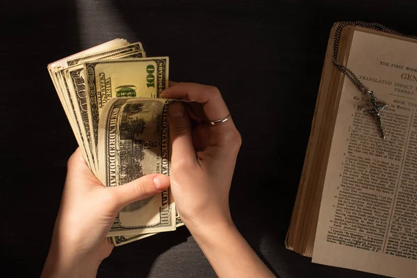 KYIV, UKRAINE - JANUARY 17, 2020: cropped view of woman counting money near holy bible with cross on dark background with sunlight — Stock Photo