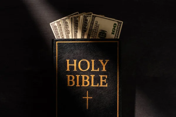Верхний вид Священной Библии с деньгами на темном фоне с солнечным светом — стоковое фото