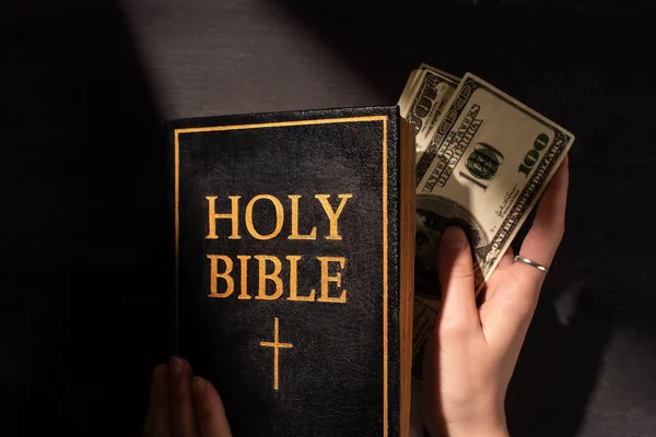 Vista recortada de la mujer sacando dinero de la sagrada biblia sobre fondo oscuro con luz solar - foto de stock
