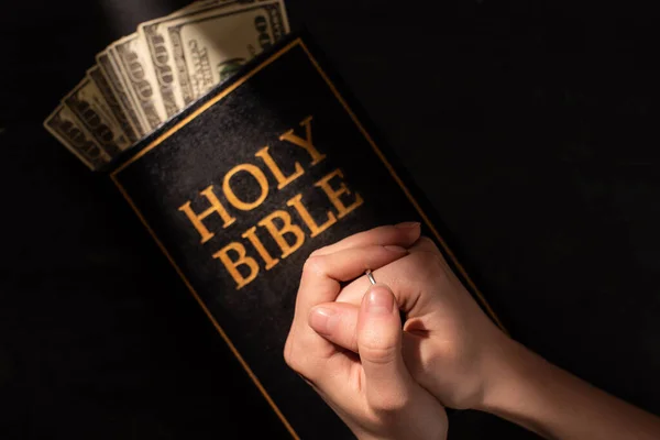 Abgeschnittene Ansicht einer betenden Frau auf heiliger Bibel mit Geld auf dunklem Hintergrund mit Sonnenlicht — Stockfoto