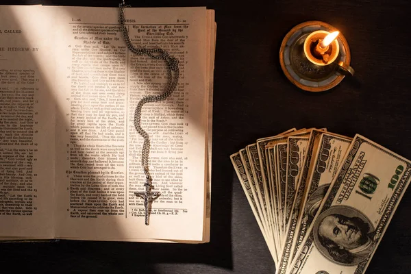 KYIV, UKRAINE - JANUARY 17, 2020: top view of holy bible with money, cross and candle on dark background with sunlight — Stock Photo