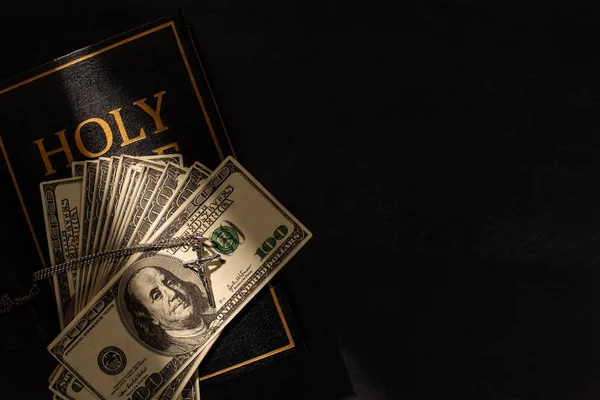 Vista superior da Bíblia sagrada com dinheiro e cruz no fundo escuro com luz solar — Fotografia de Stock