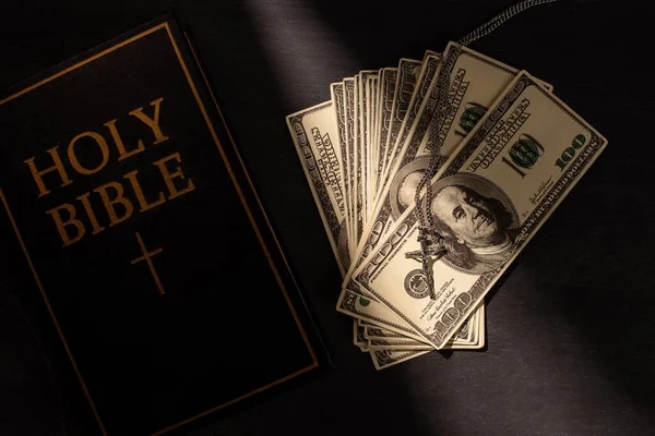 Vista superior de la sagrada biblia con dinero y cruz sobre fondo oscuro con luz solar - foto de stock