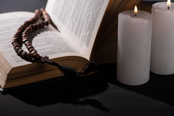 Sainte Bible avec chapelet sur fond noir foncé avec des bougies allumées — Photo de stock