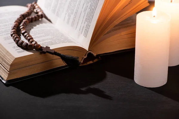 Sainte Bible avec chapelet sur fond noir foncé avec des bougies allumées — Photo de stock