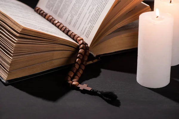 Sainte Bible avec chapelet sur fond noir foncé avec des bougies allumées — Photo de stock