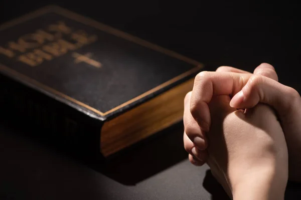 Vista recortada de la mujer orando cerca de la sagrada Biblia en la oscuridad - foto de stock