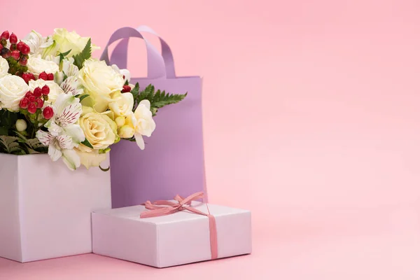 Bouquet of flowers in festive gift box with bow near violet paper bag on pink background — Stock Photo