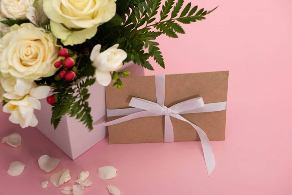 Букет цветов в праздничной подарочной коробке с открыткой и лепестками на розовом фоне — стоковое фото