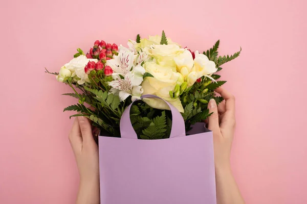 Cropped view of woman holding bouquet of flowers in violet paper bag on pink background — Stock Photo