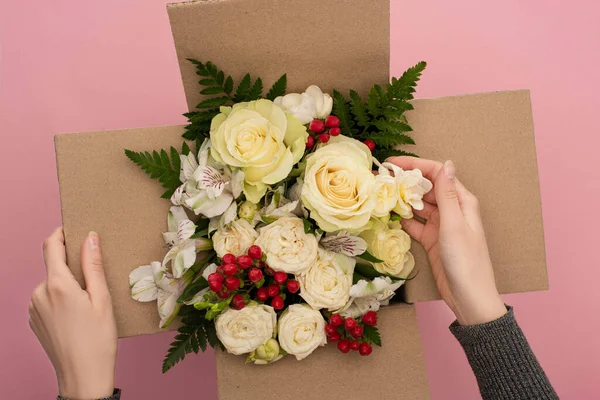 Vista parcial de mujer tocando ramo de flores en caja de cartón sobre fondo rosa - foto de stock