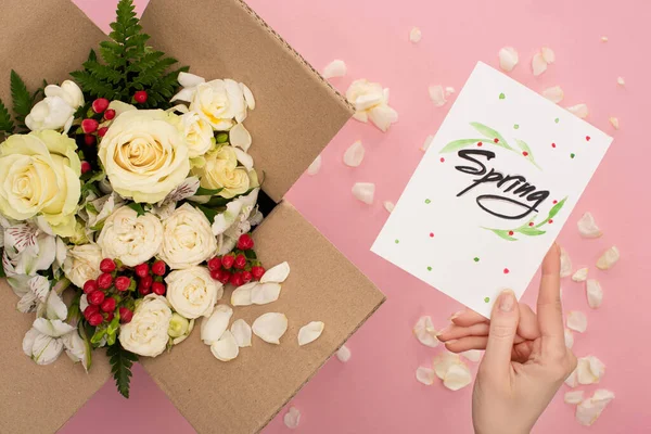Cropped view of woman holding spring card near bouquet of flowers in cardboard box on pink background — Stock Photo