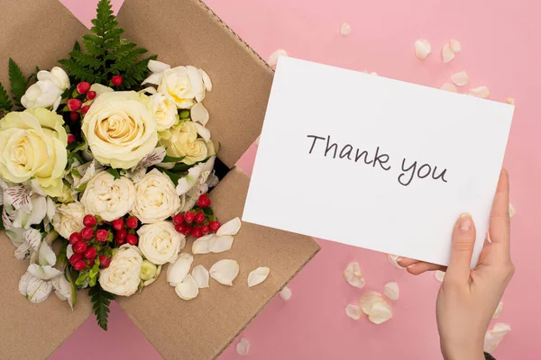 Cropped view of woman holding thank you card near bouquet of flowers in cardboard box on pink background — Stock Photo