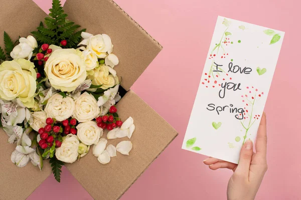 Vista recortada de la mujer sosteniendo te amo tarjeta de primavera cerca de ramo de flores en caja de cartón sobre fondo rosa - foto de stock